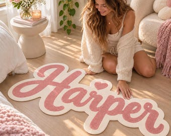 May include: A custom-shaped area rug featuring the name Harper in a textured, dusty rose pink script font against a cream-colored background. The design includes a small heart icon above the letter p, creating a soft, rounded silhouette.
