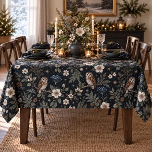 May include: A rectangular tablecloth featuring a dark navy blue background with a repeating pattern of brown owls perched on branches, white flowers, green foliage, pinecones, and gold stars. The fabric drapes over a wooden dining table.