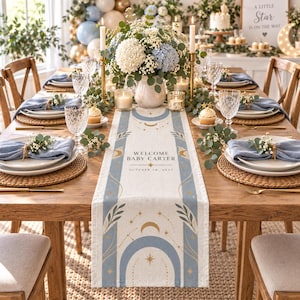 May include: A rectangular table runner featuring a celestial design with blue arches, gold stars, and moons. The center text reads "WELCOME BABY CARTER" and "OCTOBER 18, 2027". The runner is placed on a wooden dining table set with blue napkins, plates, and glasses.