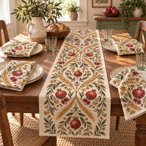 May include: A cream-coloured table runner and matching napkins featuring a repeating botanical pattern of red pomegranates, golden wheat stalks, and green leaves. The fabric has a textured appearance, and the design is symmetrical and ornate.