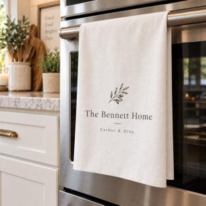 Pode incluir: Um pano de prato branco pendurado na maçaneta de um forno de aço inoxidável. O pano apresenta uma ilustração preta de um ramo de oliveira, o texto "The Bennett Home" em fonte serifada e "Gather & Dine" em uma fonte sem serifa menor.