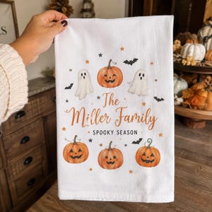 May include: A white rectangular kitchen towel featuring a printed design of four orange jack-o'-lanterns, two white ghosts, and black bats. The text reads The Miller Family and SPOOKY SEASON in orange and black script and block lettering.