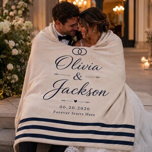 Può includere: Una coperta color crema con testo blu scuro che recita Olivia & Jackson, la data 20.06.2026 e la frase Forever Starts Here. La coperta include un grafico con due anelli intrecciati e tre strisce blu scuro sulla parte inferiore.