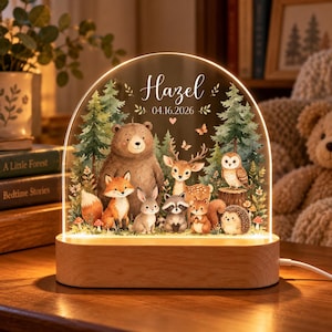 Puede incluir: Una luz nocturna de acrílico arqueada con base de madera que presenta una escena de bosque con un oso, zorro, conejo, mapache, ardilla, ciervo, búho y erizo. El texto Hazel y 04.16.2026 está impreso en la parte superior en blanco.