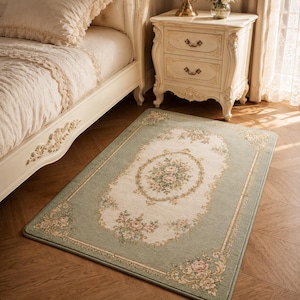 May include: A rectangular area rug featuring a sage green border and a cream-colored center. The rug displays a floral pattern with pink and green blossoms, intricate scrollwork, and decorative corner motifs in a symmetrical arrangement.