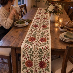 Può includere: Un lungo runner da tavola rettangolare caratterizzato da un motivo botanico ripetuto di fiori di dalia rosso scuro, foglie verdi e piccole bacche rosse su uno sfondo in tessuto beige chiaro, posizionato al centro di un tavolo in legno.