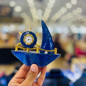 May include: A decorative desk clock shaped like a sailboat, crafted from deep blue stone. It features a gold-toned ship wheel housing the clock face with black numerals and hands, accented by a gold chain railing and a tall, pointed blue sail.