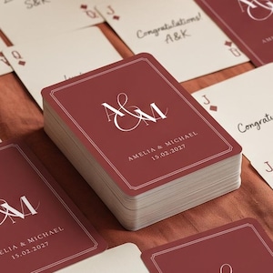 Puede incluir: Una baraja de cartas con el reverso rojo oscuro que presenta un monograma blanco de las letras A y M, los nombres Amelia & Michael y la fecha 15.02.2027. Varias cartas están extendidas, mostrando caras blancas con símbolos rojos.