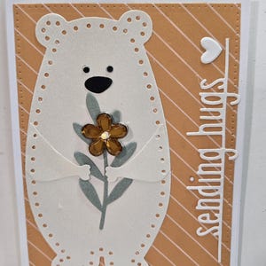 May include: A handmade greeting card featuring a white bear holding a flower with a brown center and green leaves. The bear is on a background with diagonal orange stripes. The words "sending hugs" are cut out on the right side.