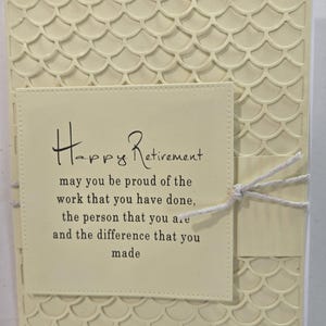 Peut inclure: Une carte de retraite cr&egrave;me avec un motif textur&eacute; en forme d'&eacute;cailles. La carte pr&eacute;sente un encart rectangulaire avec les mots "Happy Retirement" et un message sinc&egrave;re. Une ficelle blanche est nou&eacute;e autour de la carte.