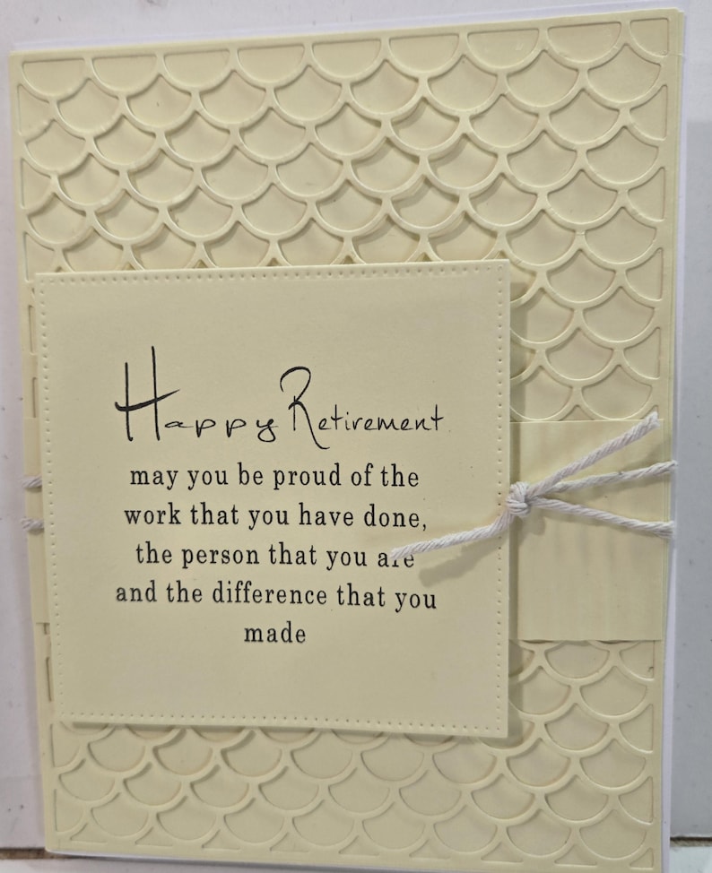 Peut inclure: Une carte de retraite cr&egrave;me avec un motif textur&eacute; en forme d'&eacute;cailles. La carte pr&eacute;sente un encart rectangulaire avec les mots "Happy Retirement" et un message sinc&egrave;re. Une ficelle blanche est nou&eacute;e autour de la carte.