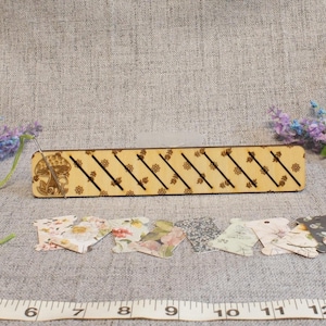May include: A rectangular light-colored wooden organizer with diagonal slots and floral engravings, accompanied by several small patterned cardboard thread bobbins. A white measuring tape shows the organizer is approximately 15 inches long.