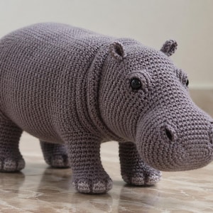 May include: A handcrafted crochet hippopotamus toy made with light purple yarn. The figure features a rounded body, four sturdy legs with detailed foot pads, small ears, and two dark, round plastic eyes set into the textured, stitched surface.