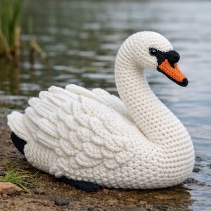 May include: A handcrafted crochet swan figurine featuring a textured white yarn body with layered wing details. The swan has a black eye, a black base at the top of its beak, and a bright orange beak. The tail feathers are tipped with black yarn.