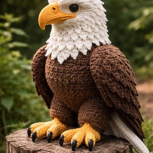 Puede incluir: Un peluche de águila calva hecho a ganchillo con cabeza blanca, cuerpo y alas de color marrón oscuro, y pico y garras de color amarillo brillante. El águila tiene ojos negros y garras oscuras, con puntadas texturizadas que crean plumas.