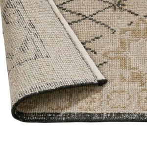 May include: A woven area rug with a folded corner revealing the underside. The rug features a beige base with a geometric pattern in dark gray and tan. The texture is visible, showing the dense pile construction and the finished edge binding.