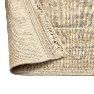 May include: A woven area rug featuring a light beige and cream base with a subtle gray geometric pattern. The rug has a folded corner revealing the underside texture and a short, fringed edge along the side, showcasing a dense, textured weave.