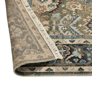May include: A woven area rug featuring a folded corner that reveals the underside. The rug displays a complex pattern in shades of sage green, beige, light blue, and black, finished with a short, light-colored fringe along the edge.