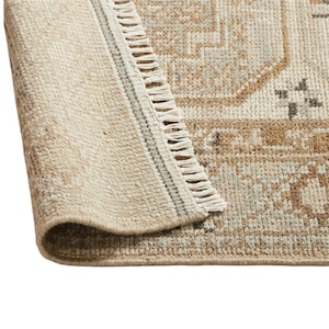 May include: A woven area rug featuring a beige and light brown color palette with a geometric pattern. The rug has a folded corner revealing a textured underside and a short white fringe along the edge, showcasing a detailed, repeating design.