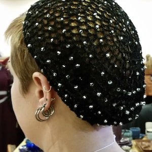May include: A black crocheted hair net, adorned with numerous small, shiny, silver-colored beads. The net covers the back of the head and is worn over short, light-colored hair. The image shows a close-up of the head and neck.