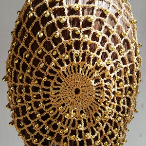 May include: A gold-colored crocheted hair net, adorned with small, golden beads. The net is designed with a circular pattern at the top and a grid-like structure throughout. The hair net is displayed on a head form, showcasing its shape and design.
