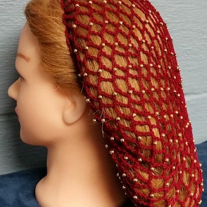 May include: A burgundy crocheted hair net with gold-colored beads. The net is designed to cover the hair and is worn on a mannequin head. The hair net is a decorative hair accessory.