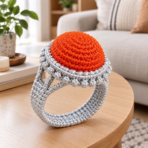 May include: A large crochet ring sculpture featuring a vibrant orange circular top set within a textured silver band. The piece is crafted with intricate yarn stitches, creating a dimensional, rounded shape that mimics the appearance of jewelry.