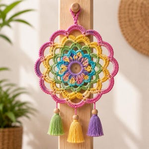 May include: A circular crochet wall hanging featuring a vibrant mandala pattern in shades of pink, yellow, green, and blue. The design includes a central ring, scalloped edges, and three hanging tassels in light green, yellow, and purple.