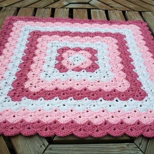 May include: A crocheted baby blanket in shades of pink, white, and burgundy. The blanket has a scalloped edge and a square pattern in the center.
