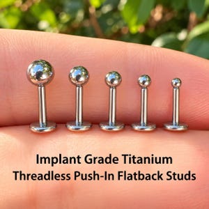 May include: Five silver-colored implant grade titanium threadless push-in flatback studs arranged in a row by size. Each stud features a smooth spherical ball top attached to a straight post and a flat circular base. Text reads: Implant Grade Titanium Threadless Push-In Flatback Studs.