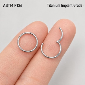 Può includere: Due anelli circolari color argento realizzati in titanio di grado implantare ASTM F136. Un anello è completamente chiuso, mentre l'altro è mostrato con il segmento incernierato parzialmente aperto, rivelando il meccanismo di chiusura.