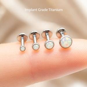 Op de afbeelding: Vier labret-piercings gemaakt van implantaat-titanium, elk voorzien van een ronde, iriserende witte opaalachtige steen in een metalen zetting. De piercings zijn op grootte gerangschikt, met de tekst Implant Grade Titanium.