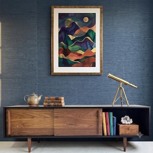May include: A framed art print featuring a stylised mountain landscape with layers of deep green, purple, and terracotta, accented by gold foil details and a golden moon, displayed above a wooden sideboard with a brass telescope and books.
