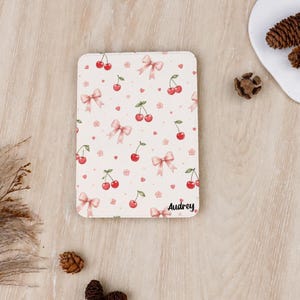 Könnte beinhalten: Eine rechteckige Tablethülle mit hellcremefarbenem Hintergrund und einem wiederkehrenden Muster aus roten Kirschen, rosa Schleifen, kleinen rosa Blüten und winzigen roten Herzen. Der Name Audrey ist unten rechts in schwarzer Schrift gedruckt.