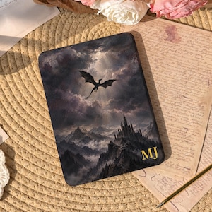 May include: A rectangular e-reader case featuring a dark, atmospheric illustration of a dragon flying over a mountain range and a castle under a cloudy sky. The bottom right corner displays the gold-colored initials MJ in a serif font.