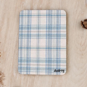 Könnte beinhalten: Ein rechteckiger Untersetzer mit einem hellblauen, weißen und beigen Karomuster. Die Oberfläche ist glatt und hat abgerundete Ecken. Der Name Audrey ist in einer schwarzen, kursiven Schriftart in der unteren rechten Ecke aufgedruckt.