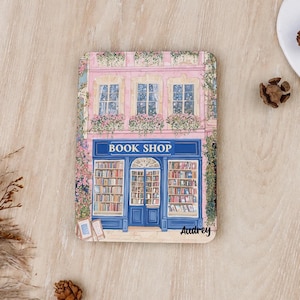 Könnte beinhalten: Ein rechteckiges Holzschild mit einer farbenfrohen Illustration eines rosa Gebäudes mit einem blauen Ladenlokal mit der Aufschrift BOOK SHOP. Die Fassade zeigt Fenster mit Blumenkästen, Kletterpflanzen und den Namen Audrey in schwarzer Schrift.