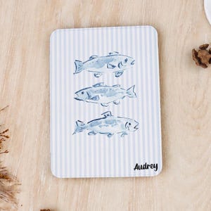 Könnte beinhalten: Eine rechteckige Platte mit hellblau-weiß gestreiftem Hintergrund, darauf drei vertikal angeordnete, aquarellartige blaue Fischillustrationen und der Name Audrey in schwarzer Schrift in der unteren rechten Ecke.