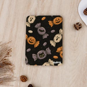 Könnte beinhalten: Ein rechteckiger Untersetzer mit schwarzem Hintergrund und einem wiederkehrenden Muster aus Halloween-Süßigkeiten, darunter orangefarbene Kürbisse, beige Geister und lila Bonbonverpackungen mit Fledermäusen und Katzen. Der Name Audrey ist in Weiß aufgedruckt.