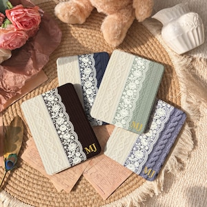 Könnte beinhalten: Vier rechteckige E-Reader-Hüllen mit vertikalem Design, bestehend aus einem cremefarbenen Zopfmuster, einem weißen Spitzenstreifen und einem einfarbigen Bereich in Dunkelbraun, Marineblau, Salbeigrün oder Lavendel, jeweils mit goldenen MJ-Initialen.