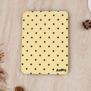 Könnte beinhalten: Eine rechteckige E-Reader-Hülle in blassem Gelb mit einem sich wiederholenden Muster aus kleinen, dunkelbraunen Sternen auf der gesamten Oberfläche. Der Name Audrey ist in schwarzer Schreibschrift in der unteren rechten Ecke aufgedruckt.