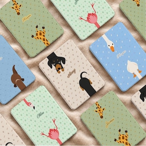 May include: A collection of rectangular e-reader cases featuring various animal illustrations including giraffes, flamingos, dachshunds, and geese. Each case displays a personalized name such as Harper, Avery, Chloe, or James printed in a script font.