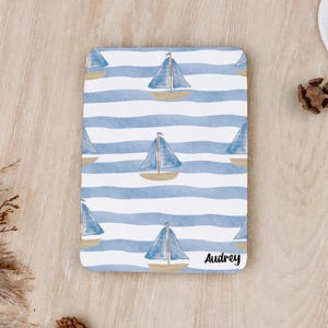 Könnte beinhalten: Ein rechteckiger Untersetzer mit einem sich wiederholenden Muster aus blauen und weißen horizontalen Wellenstreifen und Illustrationen von Segelbooten in Blau- und Beigetönen. Der Name Audrey ist unten rechts in schwarzer Schrift gedruckt.