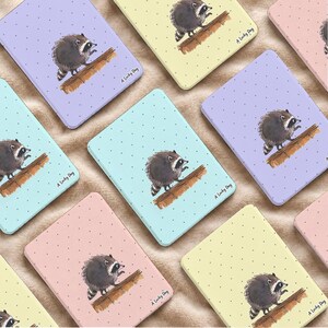 Könnte beinhalten: Eine Sammlung rechteckiger Metalldosen in Pastellfarben wie Lavendel, Hellgelb, Mintgrün und Zartrosa. Jede Dose zeigt ein Punktmuster und eine Aquarellillustration eines Waschbären auf einem Holzstamm mit dem Schriftzug A Lucky Day.