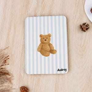 Könnte beinhalten: Eine rechteckige Platte mit einem hellblau-weißen vertikalen Streifenmuster. In der Mitte der Platte befindet sich eine Illustration eines braunen Teddybären. Der Name Audrey ist in schwarzer Schreibschrift in der unteren rechten Ecke gedruckt.