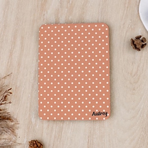 Könnte beinhalten: Eine rechteckige E-Reader-Hülle mit terrakottafarbenem Hintergrund und einem wiederkehrenden Muster aus kleinen weißen Punkten. Der Name Audrey ist in einer schwarzen Schreibschrift in der unteren rechten Ecke der Hülle aufgedruckt.