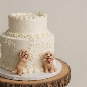 May include: A two-tier white cake decorated with piped floral patterns and small white pearls. Two light brown dog figurines with pink collars are positioned at the base of the cake, one standing on its hind legs and the other sitting down.