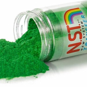 Op de afbeelding: Een doorzichtige plastic pot ligt op zijn kant en morst felgroen poeder. Het etiket toont een regenboogafbeelding boven de rode tekst N.S.T. en de blauwe tekst NEW SYSTEM TRANSFER. Het fijne, korrelige poeder heeft een verzadigde groene kleur.