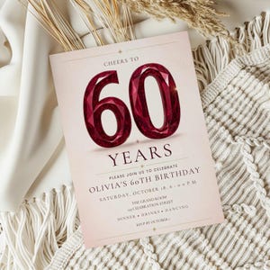 May include: A rectangular invitation card with a pale pink background featuring the text CHEERS TO 60 YEARS in dark red, with the number 60 rendered in a faceted ruby-like style. The card includes details for Olivia's 60th birthday celebration.