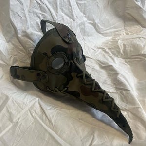 Op de afbeelding: Een pestmeestermasker met een camouflagepatroon in tinten groen, bruin en zwart. Het masker heeft een lange, puntige snavel, ronde oogopeningen met doorzichtige glazen en een dikke groene koordsluiting langs de middennaad en zijkanten.